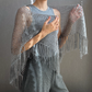 A person wearing a gray open-knit fringed shawl or poncho over a ribbed gray top and matching gray pants, posed against a plain gray wall.