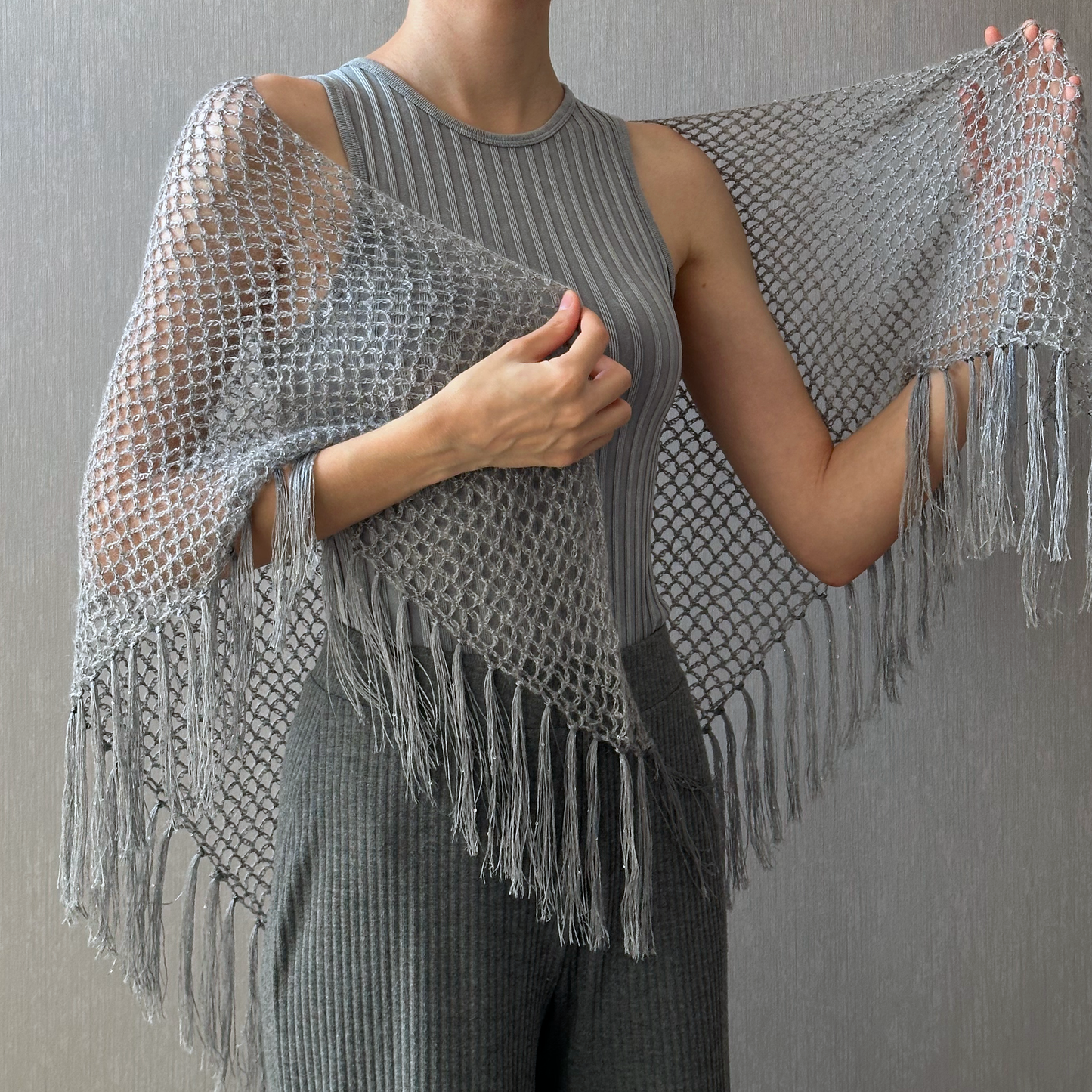 A person wearing a gray sleeveless ribbed top and matching gray pants, draped in a large open-knit fringed shawl or poncho over the shoulders, against a textured gray wall.