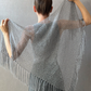 A woman shown from behind wearing and holding out a large open-front knit shawl with fringe, in a light gray color against a textured gray wall.