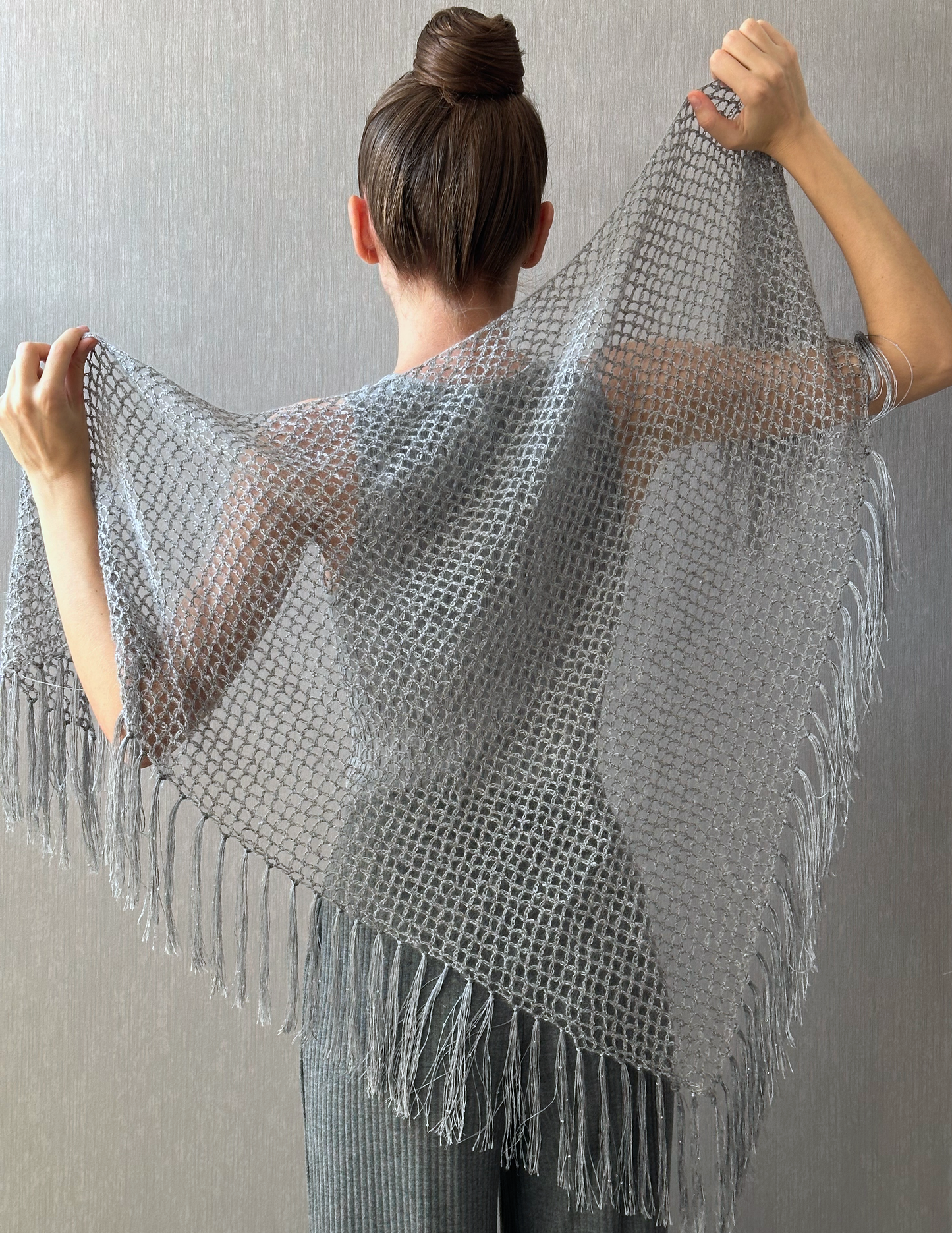 A woman shown from behind wearing and holding out a large open-front knit shawl with fringe, in a light gray color against a textured gray wall.