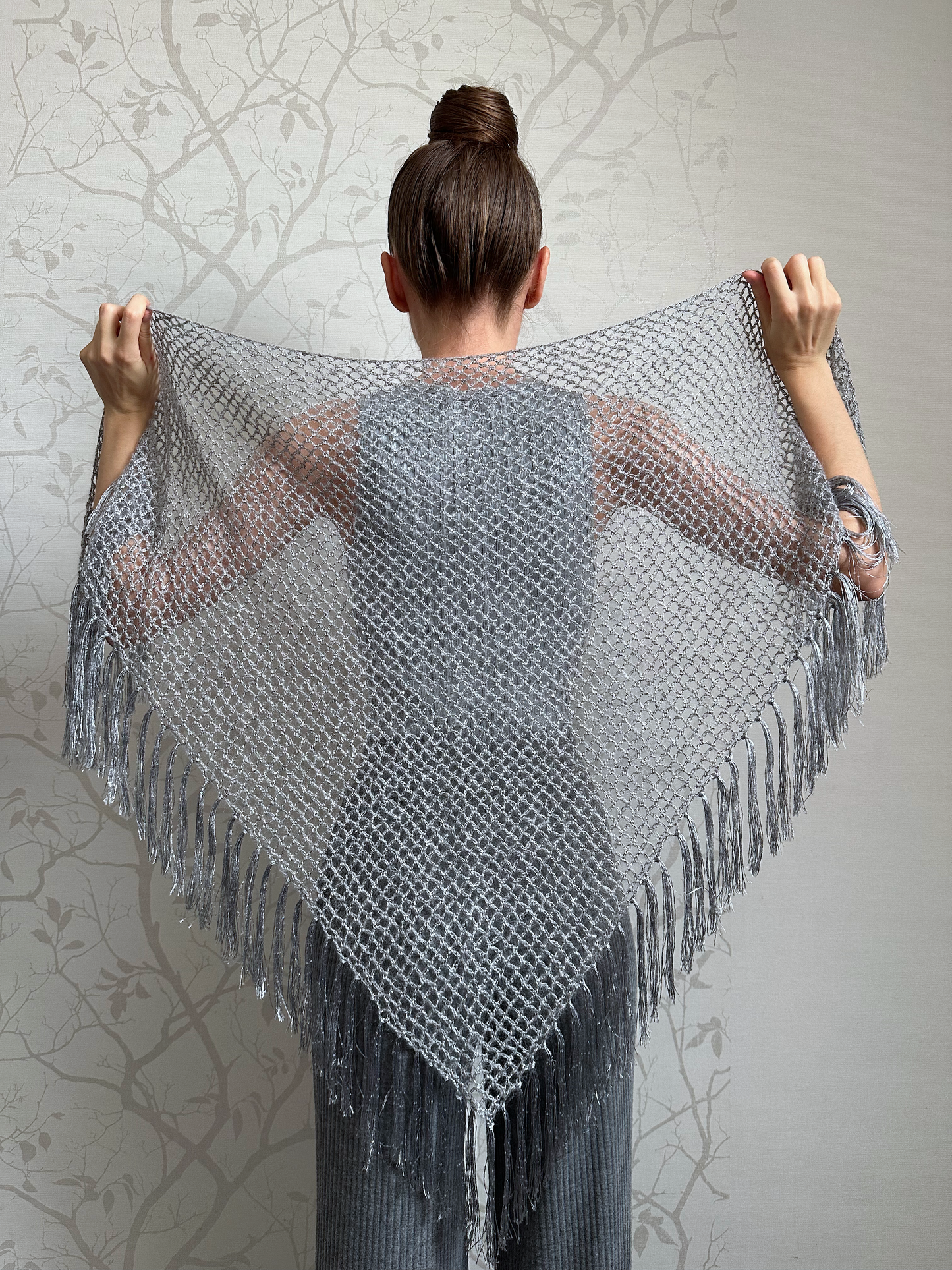 A woman shown from behind wearing and holding up a large triangular gray knit shawl with fringe, against a light gray patterned wall.