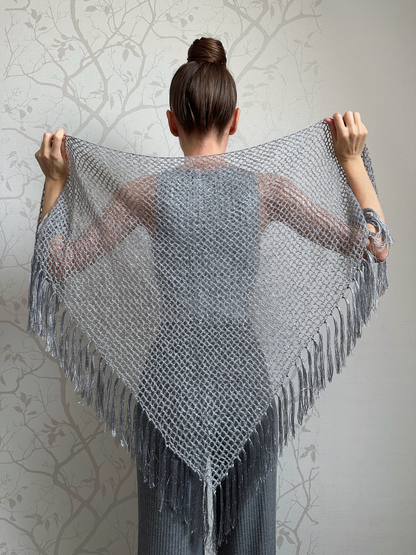 A woman shown from behind wearing and holding up a large triangular gray knit shawl with fringe, against a light gray patterned wall.