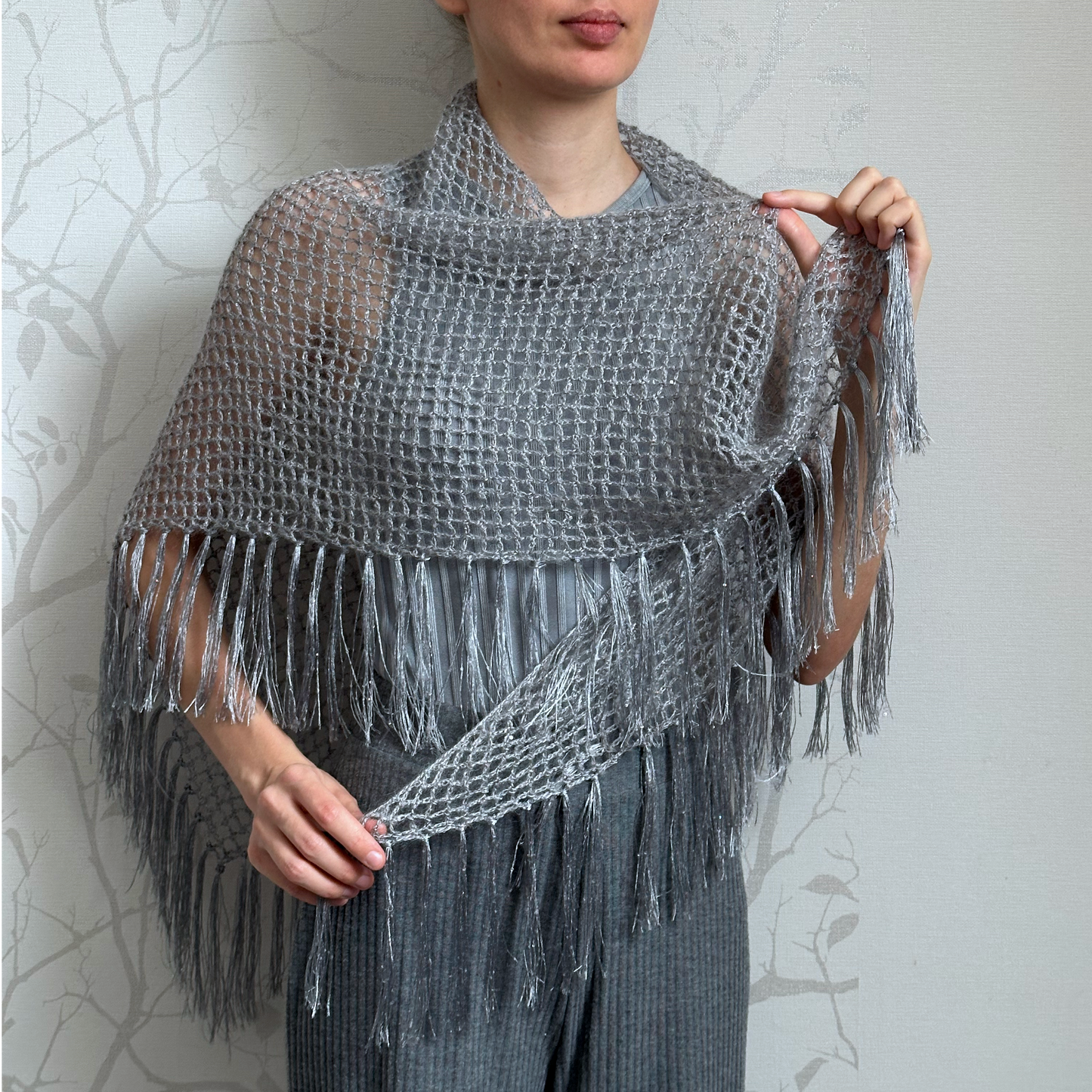 A woman models a gray open-knit fringed shawl over her shoulders and around her arms, holding the edges to show the texture and drape. The background is a light gray wall with a faint tree branch pattern.