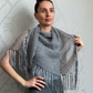 A woman wearing a gray open-knit fringed shawl over a dark gray ribbed top and trousers, standing against a light gray patterned wallpaper background.
