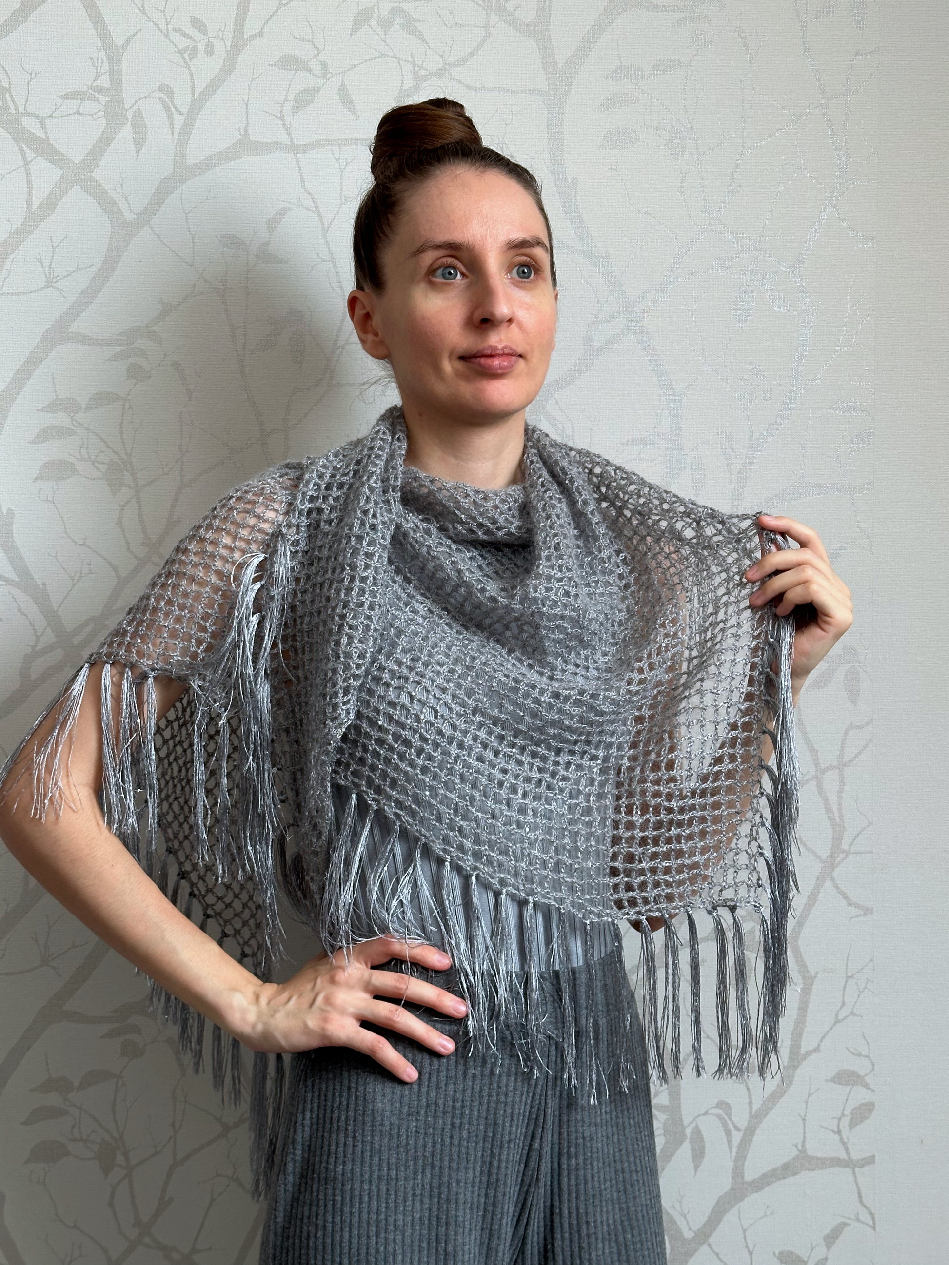 A woman wearing a gray open-knit fringed shawl over a dark gray ribbed top and trousers, standing against a light gray patterned wallpaper background.