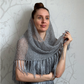A woman wearing a chunky gray knit shawl with fringe, posing against a light gray patterned wall.