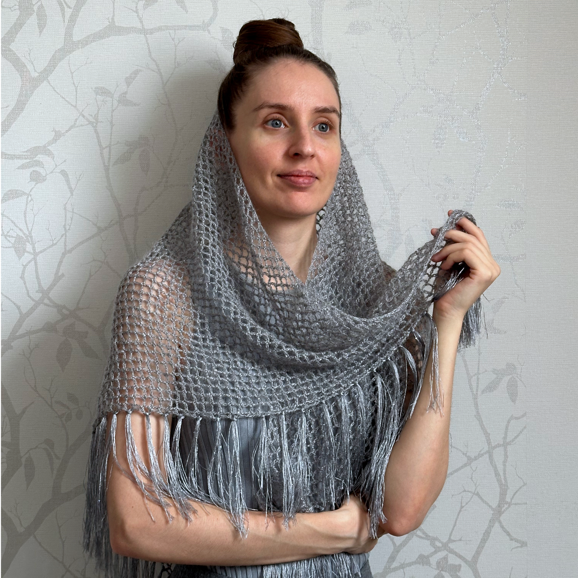 A woman wearing a chunky gray knit shawl with fringe, posing against a light gray patterned wall.