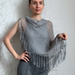 A woman modeling a gray open-knit fringed shawl draped over her shoulders, paired with gray knit pants, standing against a light gray patterned wall.