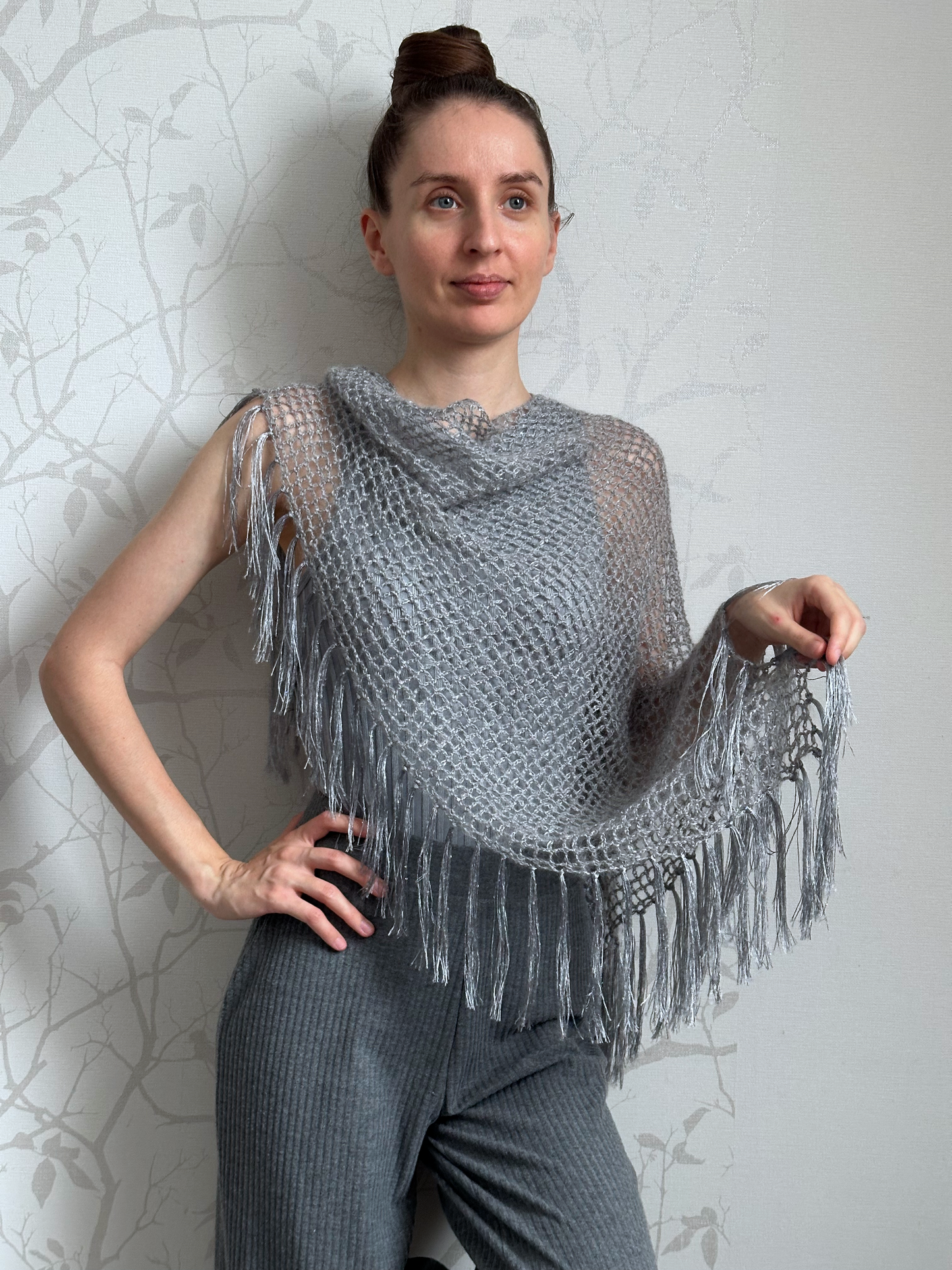 A woman modeling a gray open-knit fringed shawl draped over her shoulders, paired with gray knit pants, standing against a light gray patterned wall.