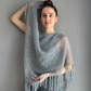 A woman modeling a light gray open-knit fringed shawl or poncho against a textured gray wall.