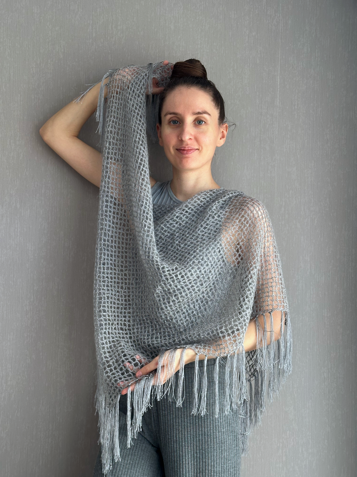 A woman modeling a light gray open-knit fringed shawl or poncho against a textured gray wall.