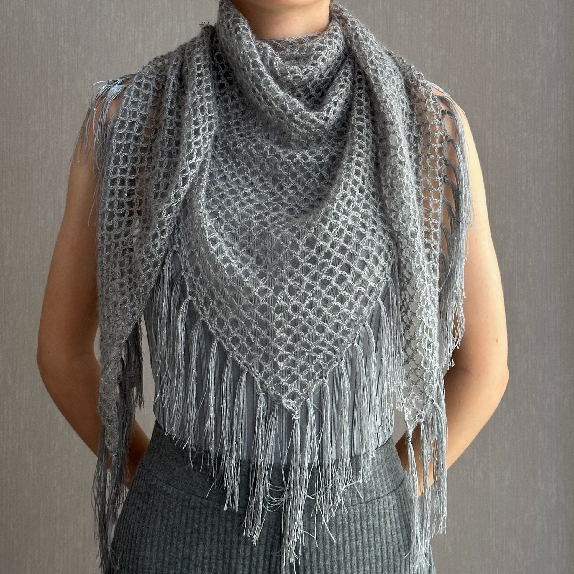 A gray open-knit cowl neck scarf with long fringe tassels, draped over a person's shoulders and torso against a neutral gray background.