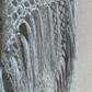 Close-up of a light gray knitted textile with long tassel fringe, shown against a neutral beige background.