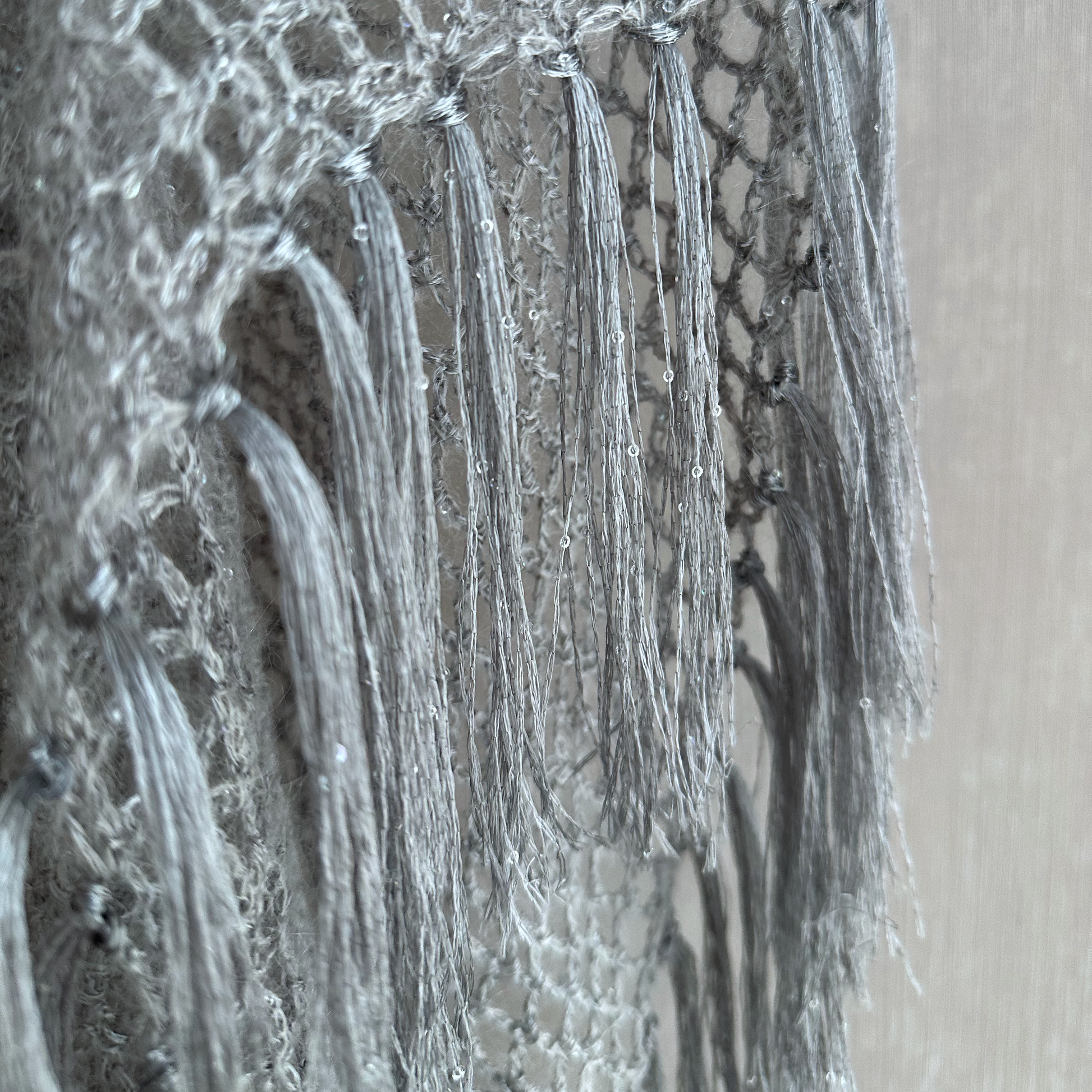 Close-up of a light gray knitted textile with long tassel fringe, shown against a neutral beige background.