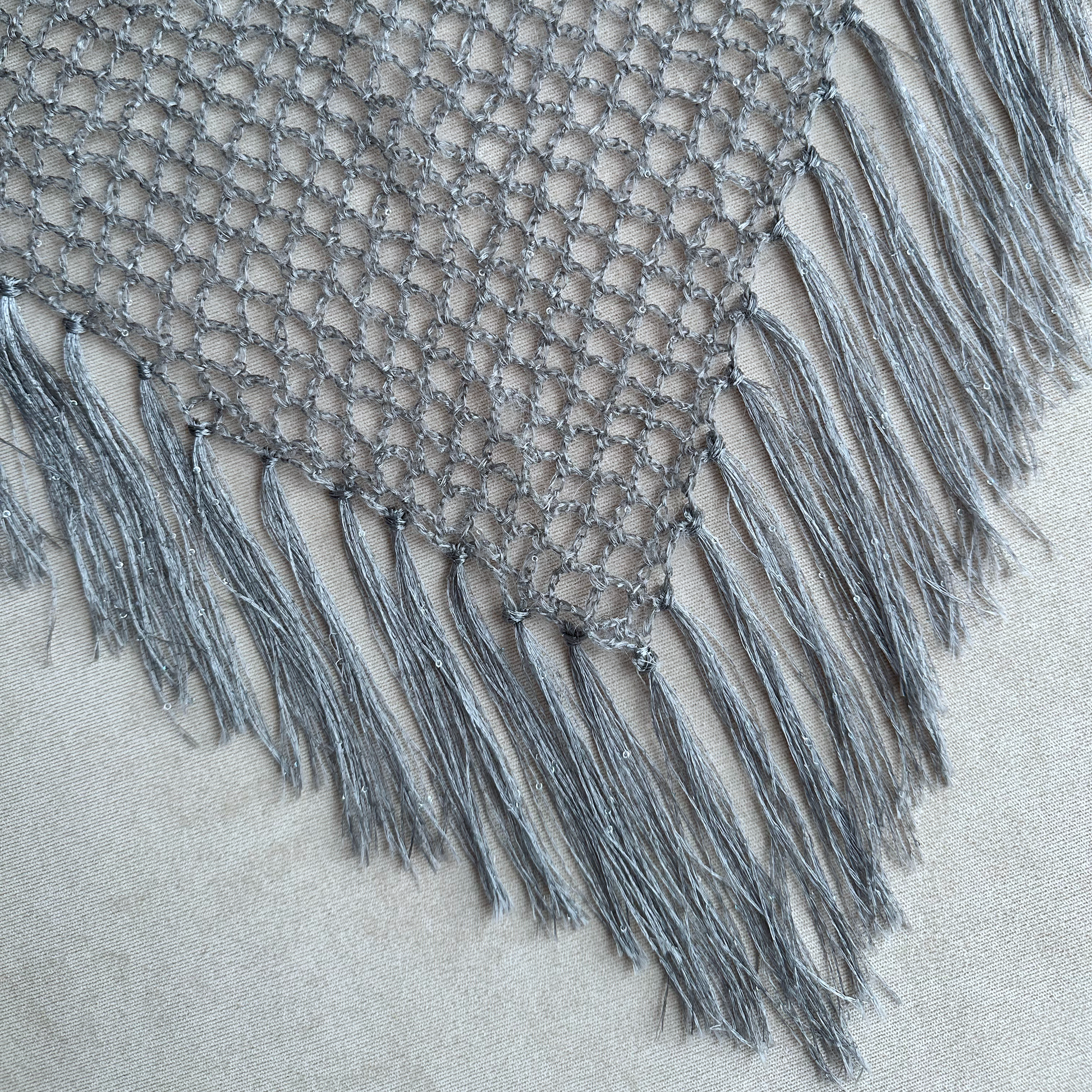 A close-up of a gray crocheted or knitted textile with an open mesh pattern and long fringe tassels, laid on a light beige fabric background.