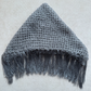 A gray open-knit triangular head covering with fringe along the bottom edge, laid flat on a light beige textured background.