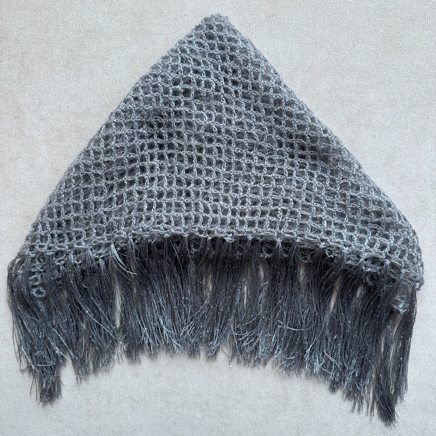 A gray open-knit triangular head covering with fringe along the bottom edge, laid flat on a light beige textured background.
